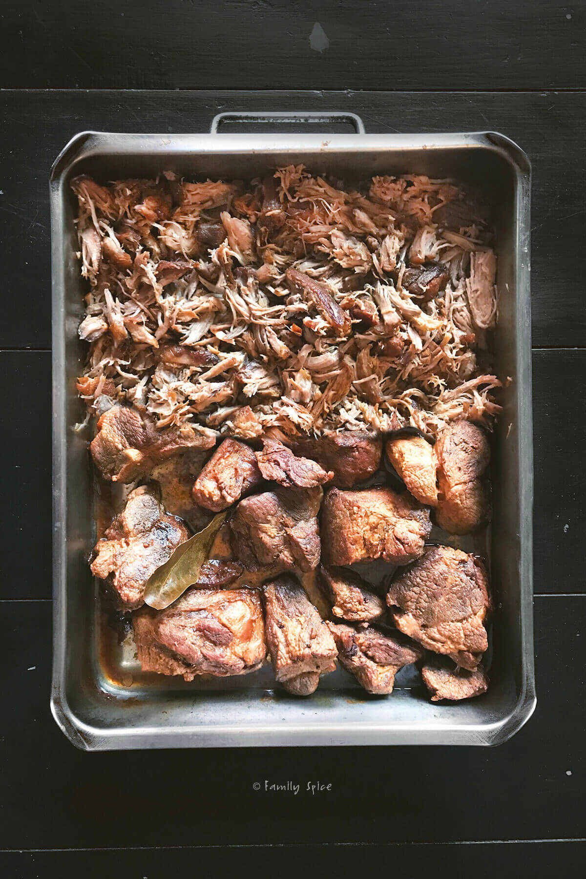 Overhead view of a roasting pan half filled with roasted pork shoulder and half filled with shredded pulled pork