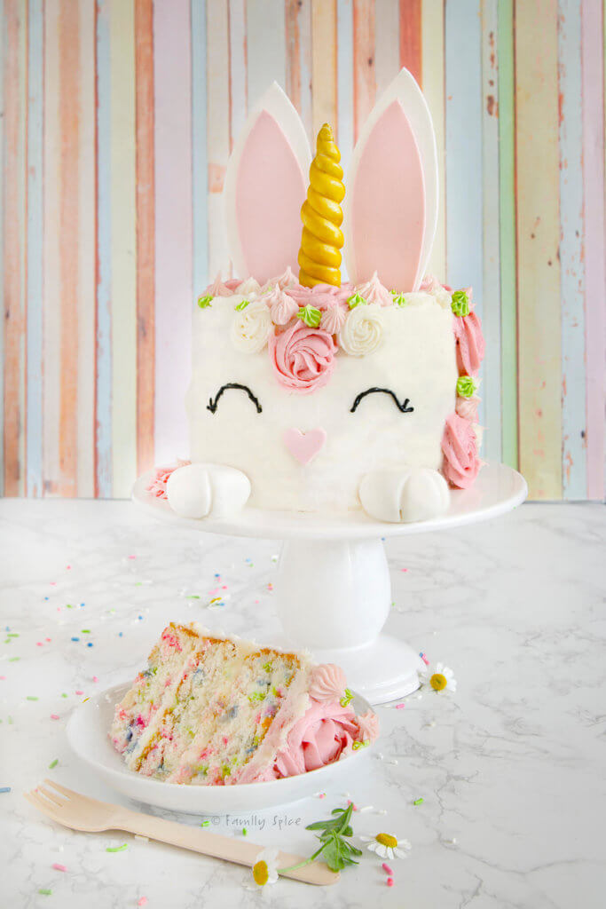 A bunny unicorn cake in pastel pinks and greens with a slice of funfetti cake on a white plate next to it
