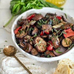 Closeup of a white bowl with roasted vegetable salad with basil and olive oil behind it.
