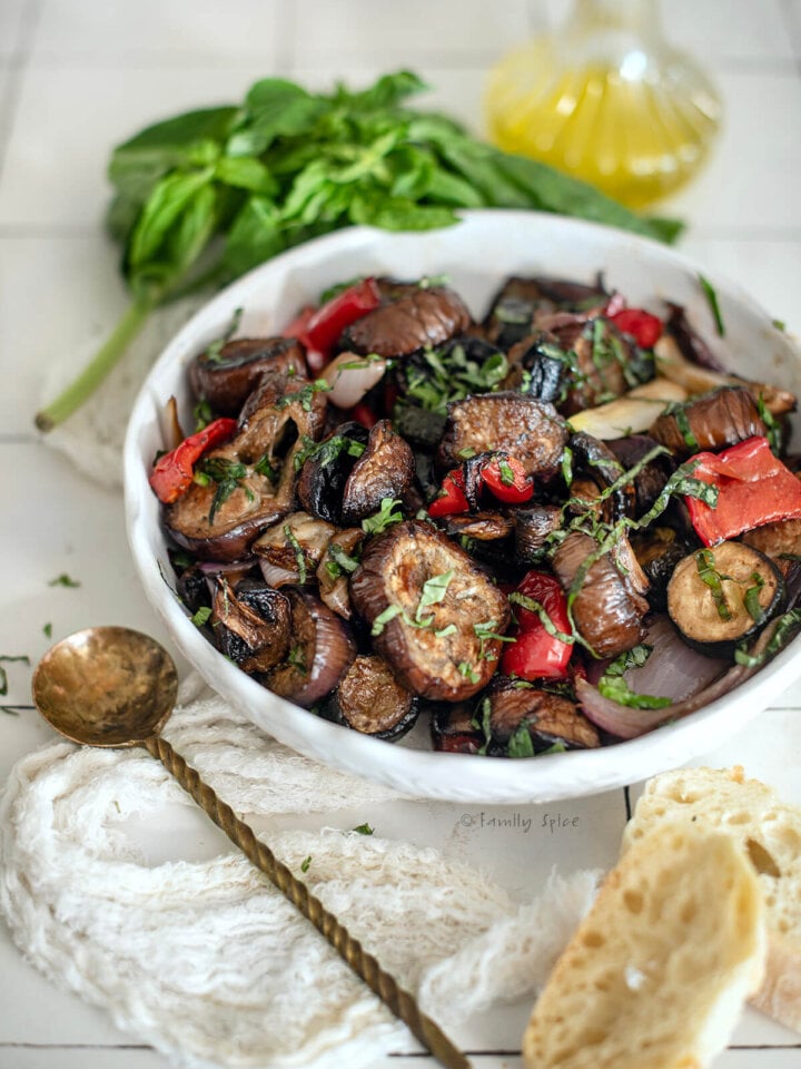 Closeup of a white bowl with roasted vegetable salad with basil and olive oil behind it.
