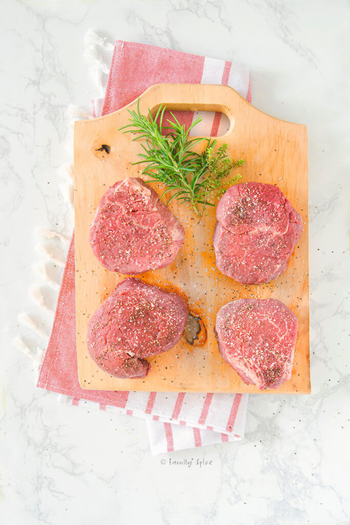 Top view of 4 raw and seasoned filet mignons on a wood cutting board with fresh herbs