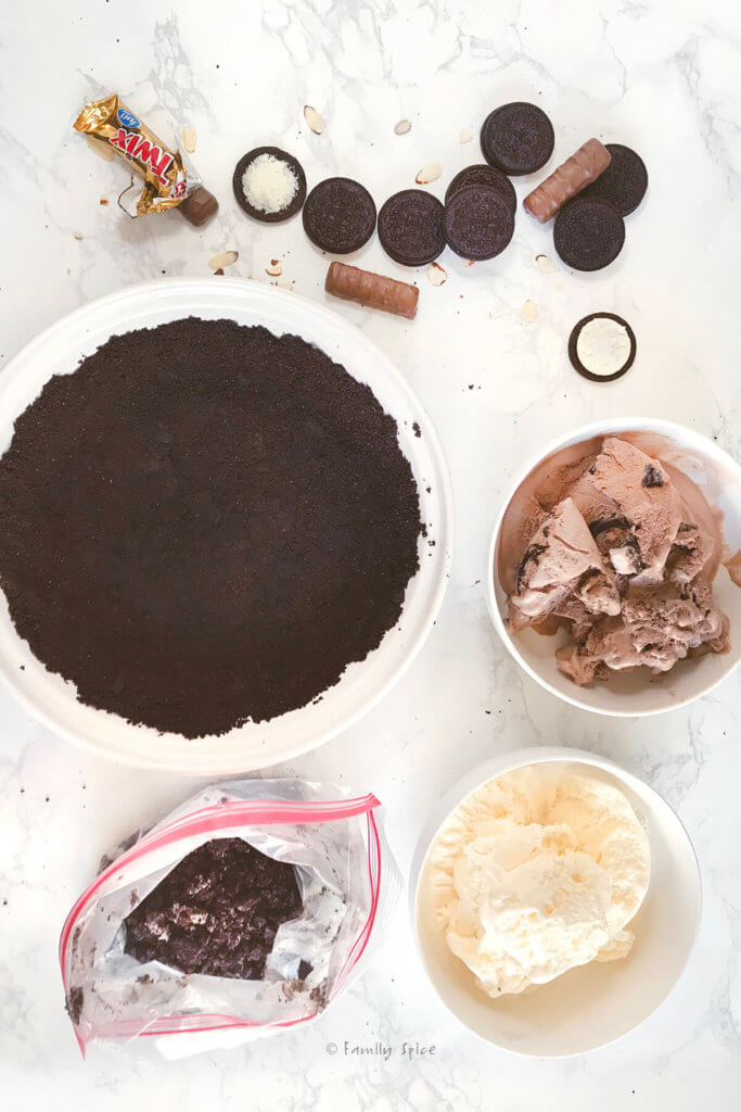 Oreo crust pressed into a white pie dish with bowls of ice cream and chocolate candies next to it
