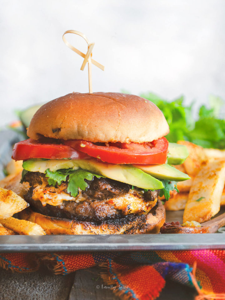Side view of a queso stuffed burger with tomatoes, cilantro and avocado slices on a tray with fat steakhouse french fries