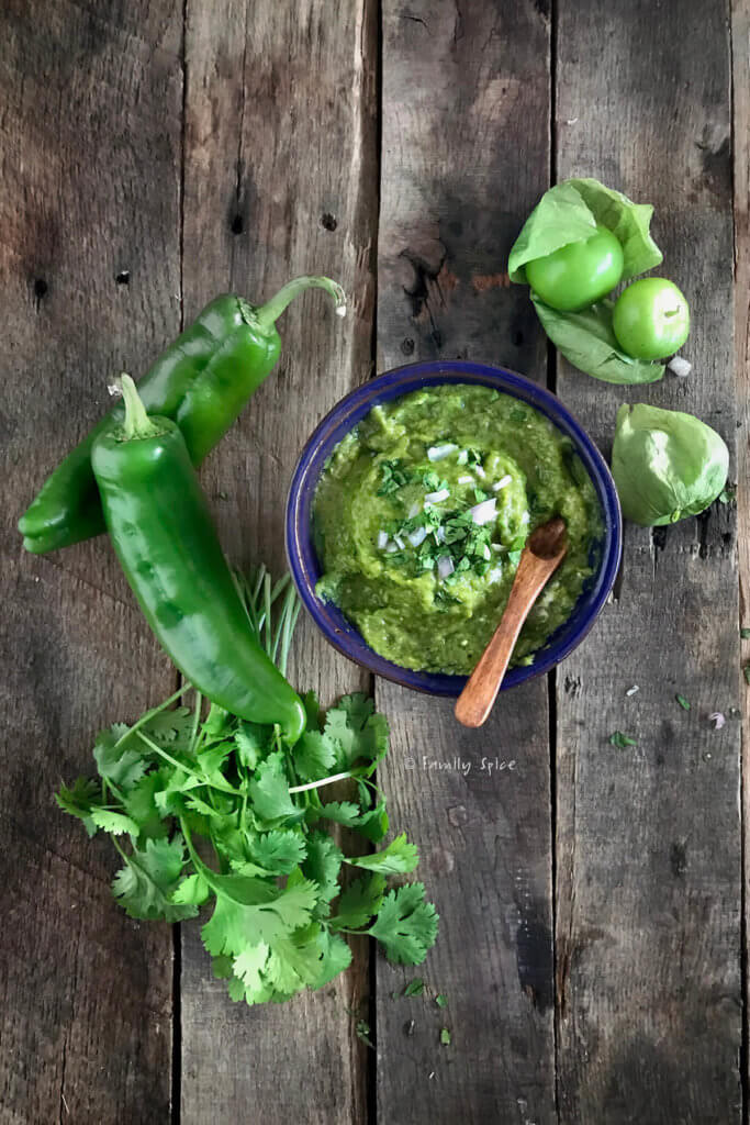 A bowl of chili verde sauce with cilantro, green chiles and tomatillos next to it