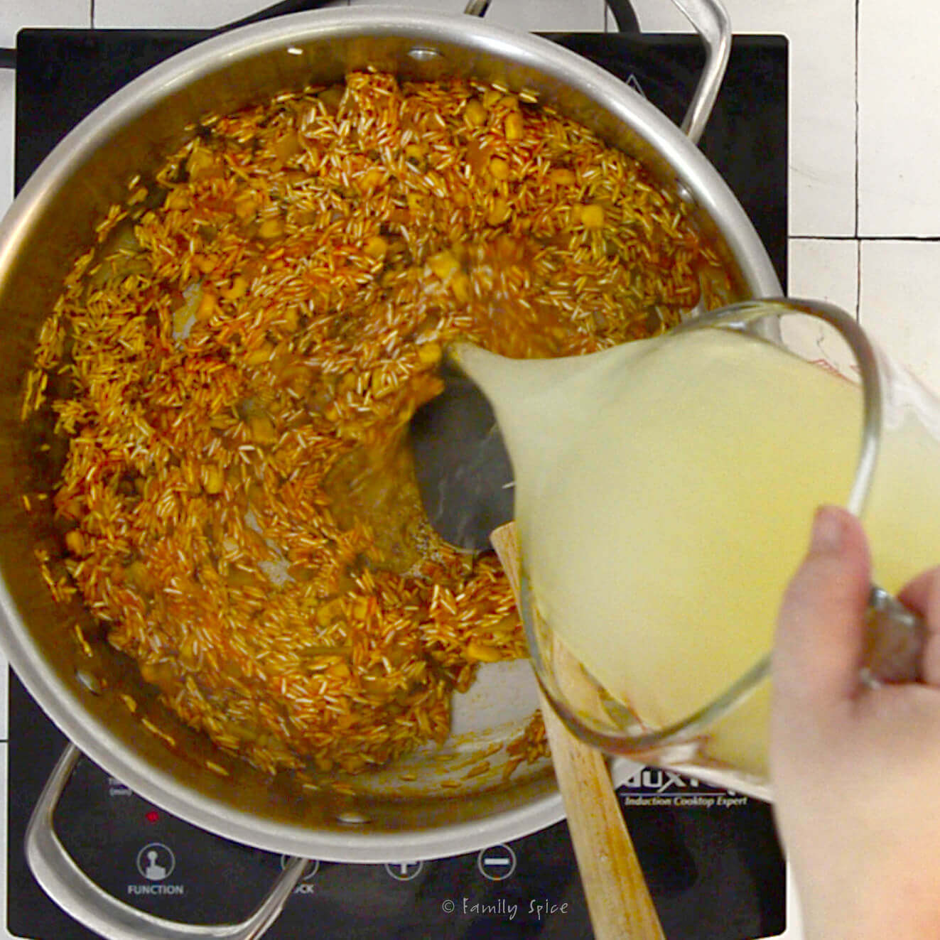 A hand pouring chicken broth into a stainless pot with remaining ingredients cooking to make spanish rice