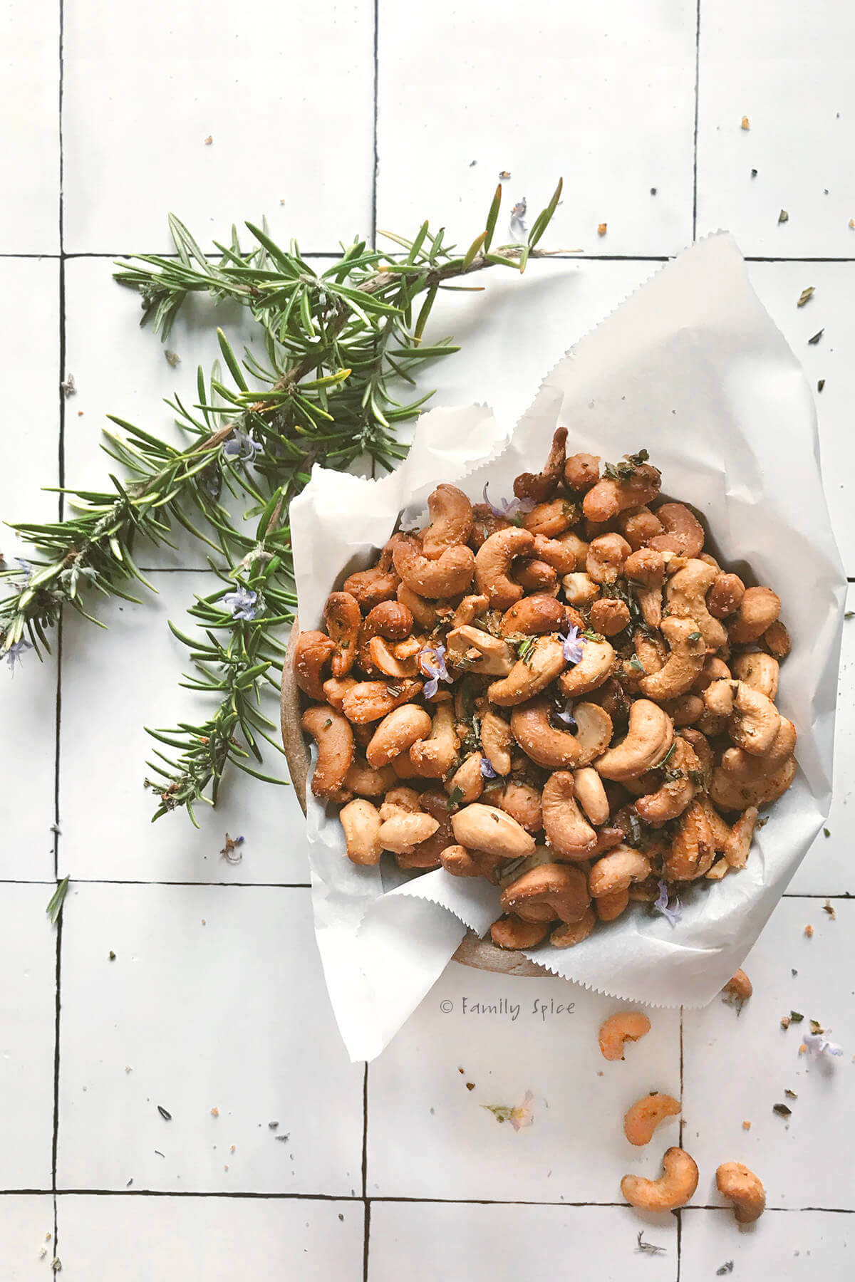 A bowl of rosemary roast cashews in a bowl lined with parchment paper with branches of rosemary next to it