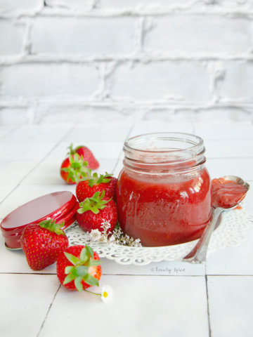 A bottle of strawberry rhubarb jam surrounded by fresh strawberries and white flowers with a spoon of jam next to it