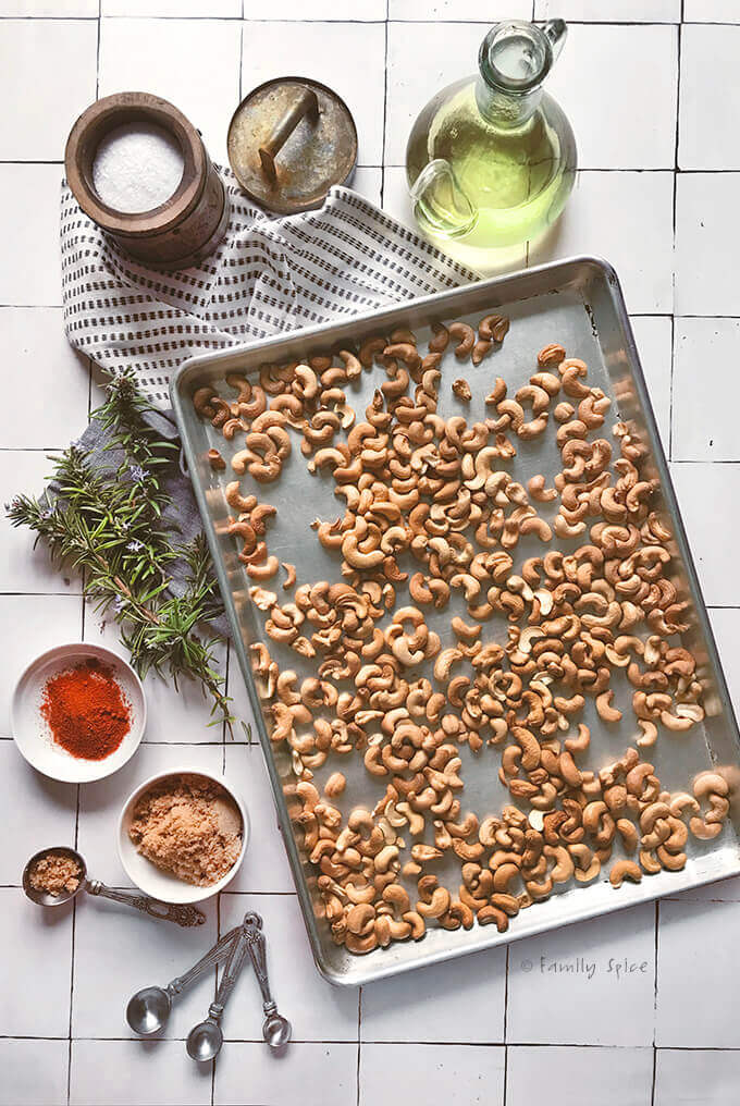 Roasted cashews in a baking sheet surrounded by rosemary, oil and seasonings