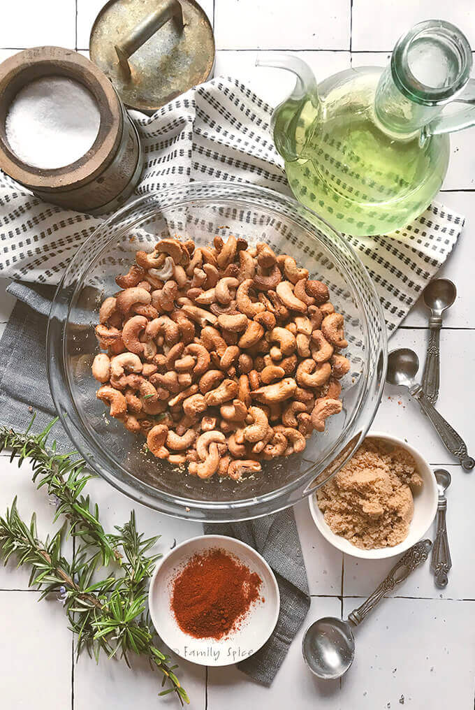 A bowl of rosemary roast cashews in a bowl surrounded by ingredients used to make it