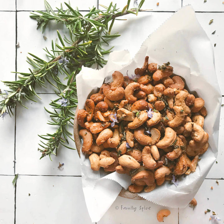 A bowl of rosemary roast cashews in a bowl lined with parchment paper with branches of rosemary next to it