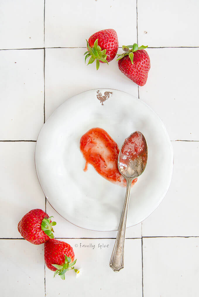 A white plate with strawberry rhubarb jam smeared on it and jam on a spoon, surrounded by some fresh strawberries.