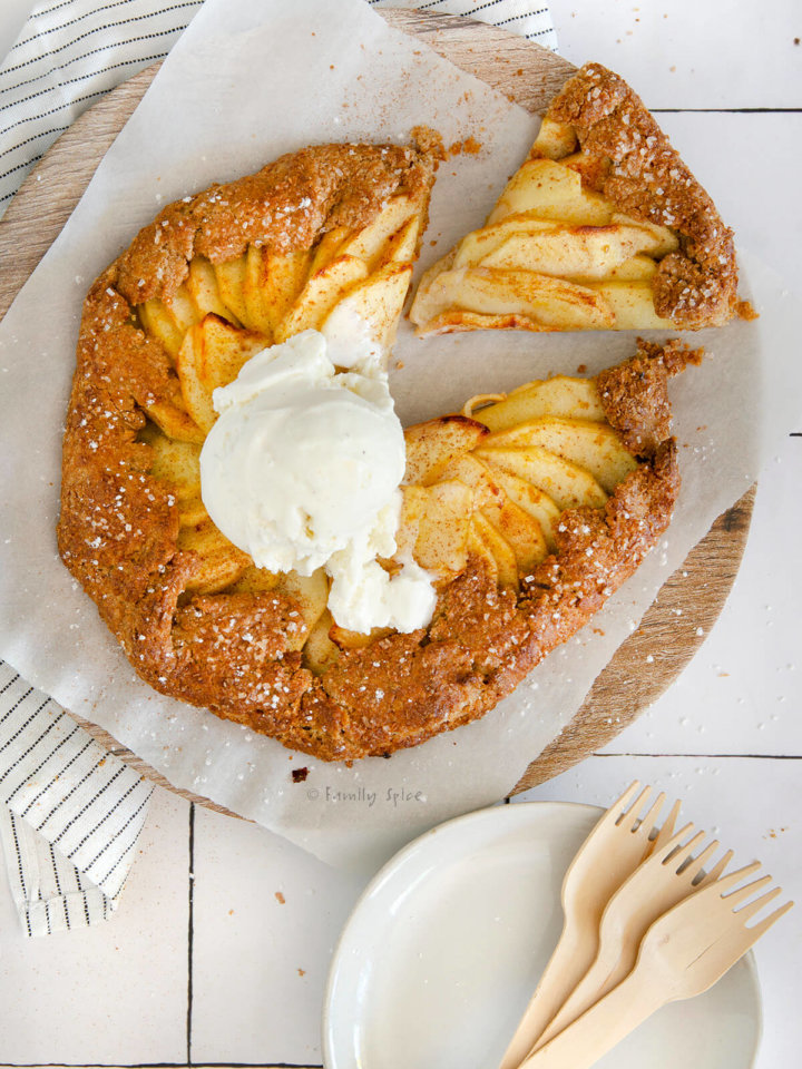 A freshly baked whole wheat apple galette topped with vanilla ice cream with a small slice cut out and small white plates and bamboo forks next to it