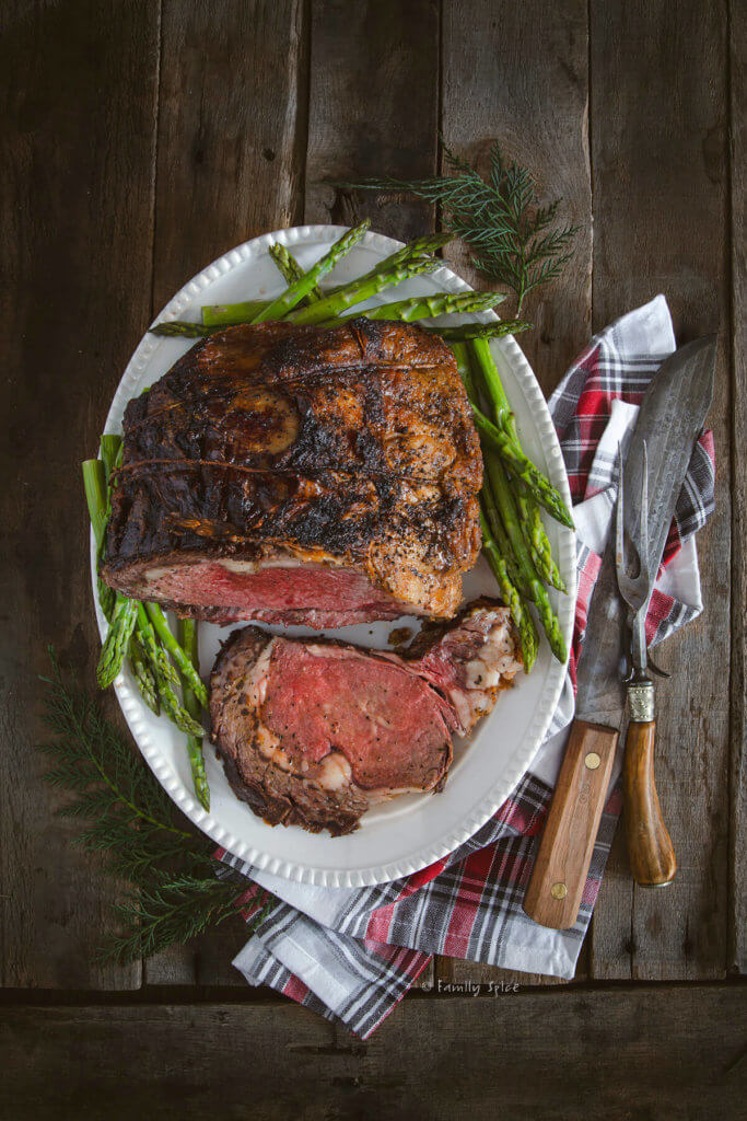A prime rib roast with a slice cut out on a white serving plate surrounded with steamed asparagus