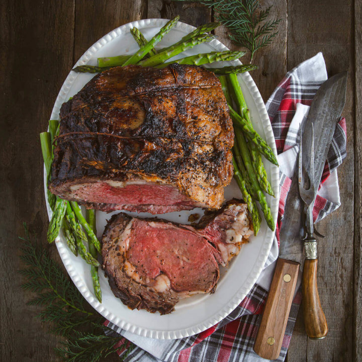 A prime rib roast with a slice cut out on a white serving plate surrounded with steamed asparagus