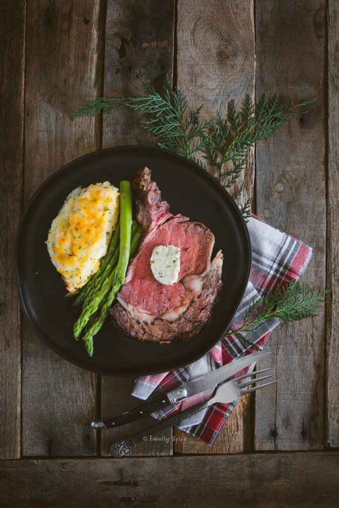A black dinner plate with a slice of ribeye with a pat of compound butter, steamed asparagus and twice baked potato
