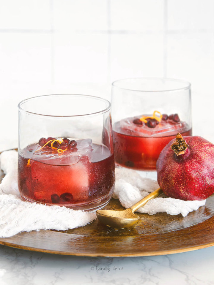 Sideview of two glasses with a large ice cube and pomegranate gin sling inside garnished with pomegranate and orange zest on a golden tray