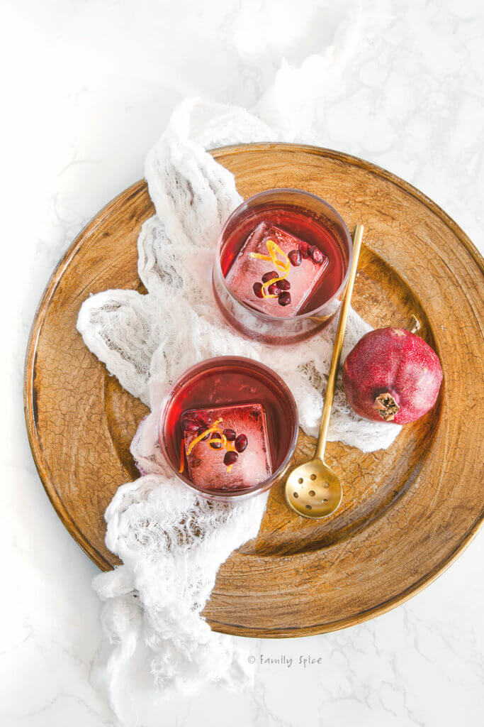 Overhead shot of two glasses with a large ice cube and pomegranate gin sling inside garnished with pomegranate and orange zest on a golden tray
