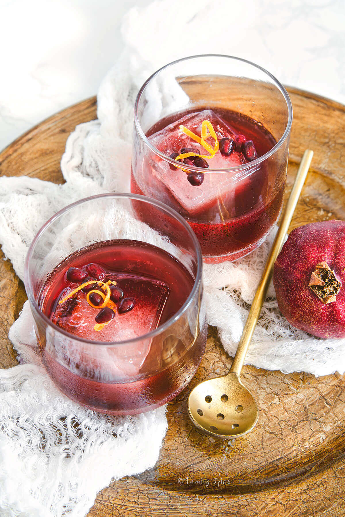 Closeup on two glasses with a large ice cube and pomegranate gin sling inside garnished with pomegranate and orange zest on a golden tray