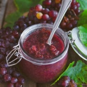 Closeup of a jar of grape jam with a spoon in it surrounded by red grapes and grape leaves