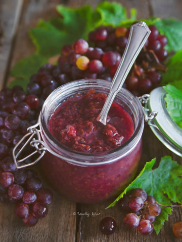Closeup of a jar of grape jam with a spoon in it surrounded by red grapes and grape leaves