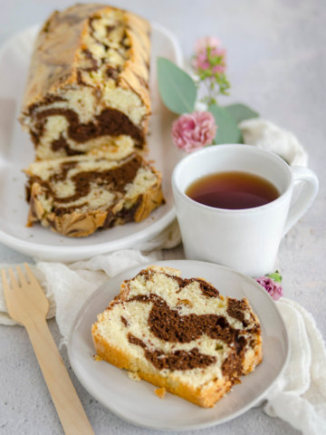 Close up of a slice of marble loaf cake on a white plate with a mug of tea and the remaining cake behind it