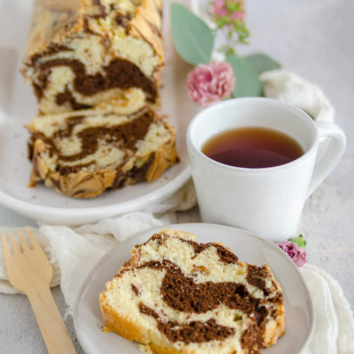 Close up of a slice of marble loaf cake on a white plate with a mug of tea and the remaining cake behind it