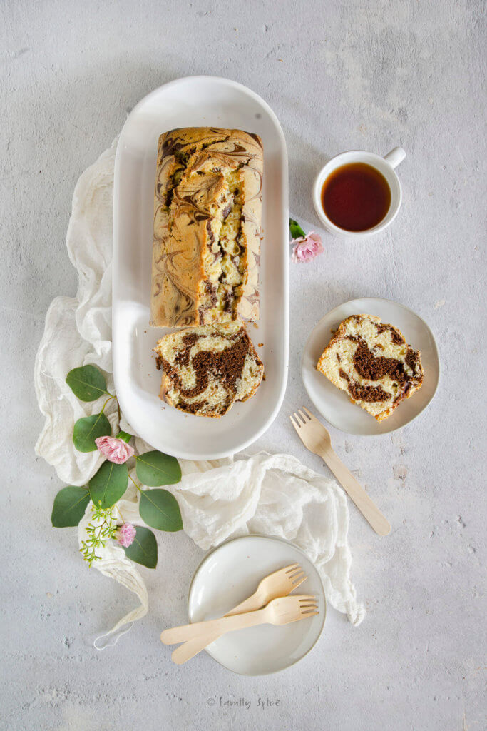 Top view of a marble loaf cake on a white platter with a slice on a plate next to it, several white plates and a mug of tea