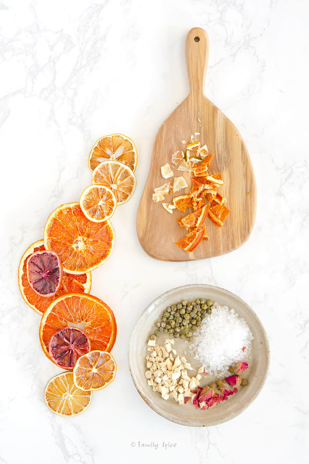 A cutting board with chopped dried oranges, a shallow bowl with coarse sea salt, green pepper corns, dried ginger and dried roses with assorted dried citrus slices