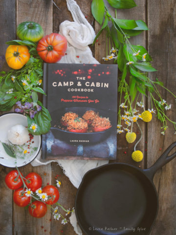 The Camp & Cabin Cookbook by Laura Bashar with fresh ingredients, flowers and cast iron pan around it