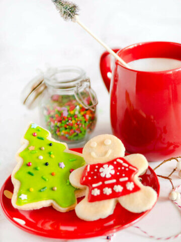 A red plate with a couple decorated Christmas sugar cookies and red mug with milk in it