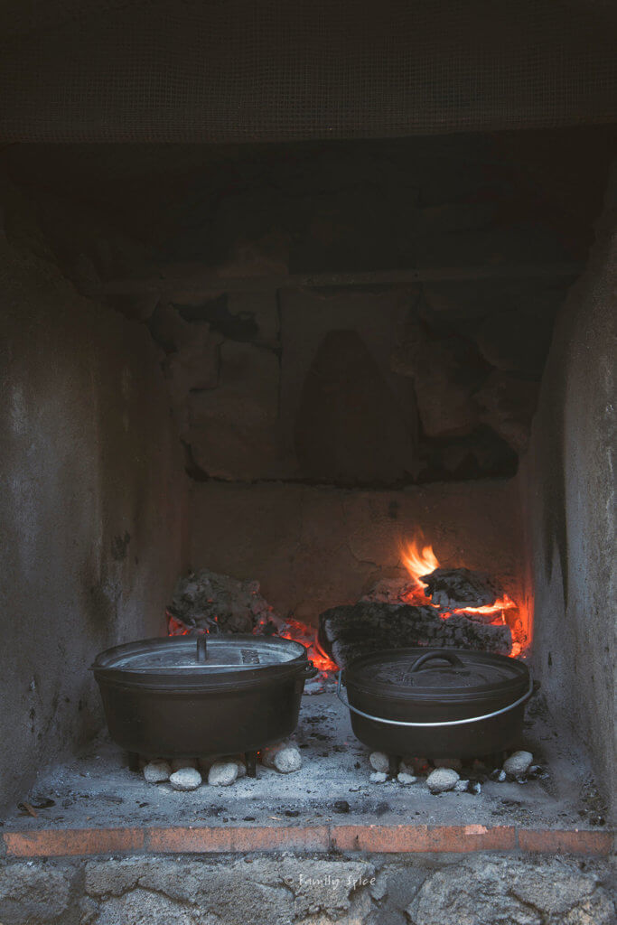 Two Dutch ovens with coals underneath in a big fire hearth with fire behind it