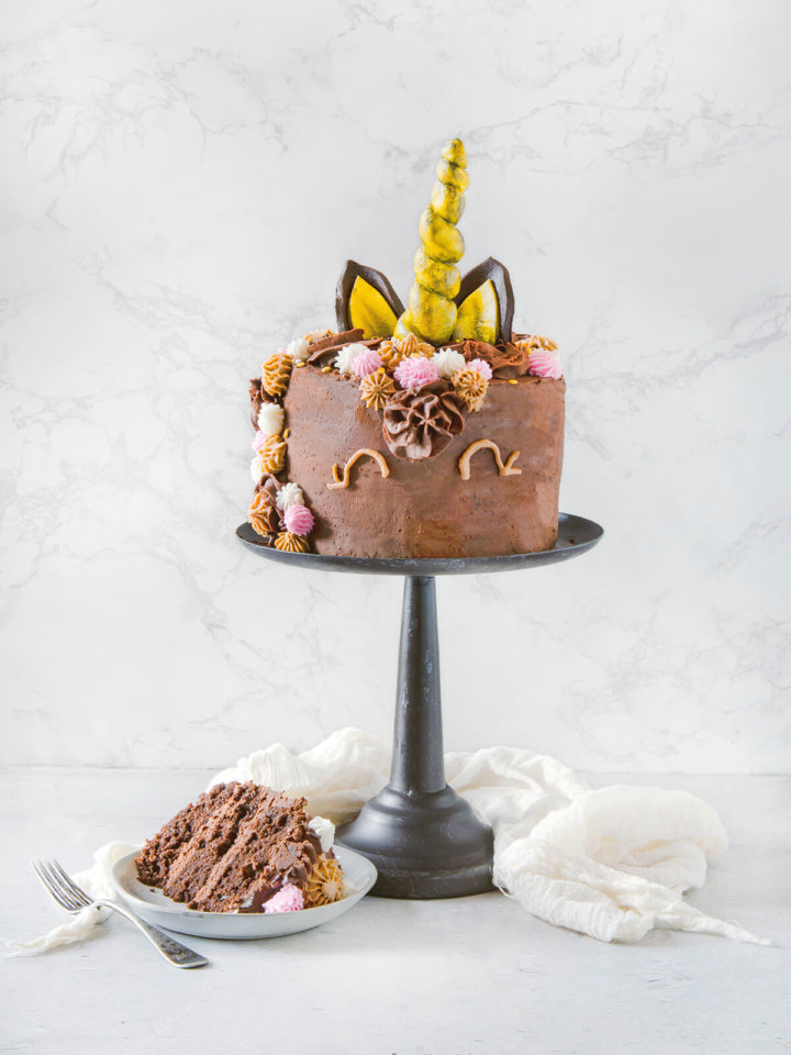 A slice of chocolate cake next to a chocolate unicorn cake on a metal cake stand