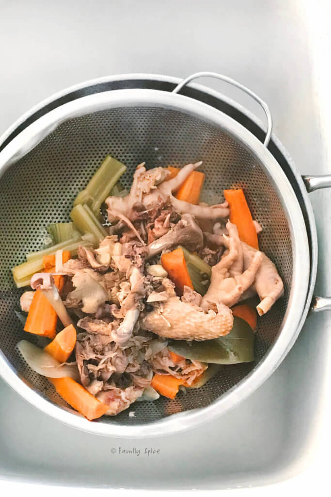 A metal strainer in a white kitchen sink with cooked chicken feet and vegetables that made chicken bone broth