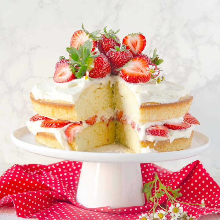 A vanilla cake frosted minimally with strawberry slices in between, with a slice cut out and topped with more fresh strawberries and chamomile flowers