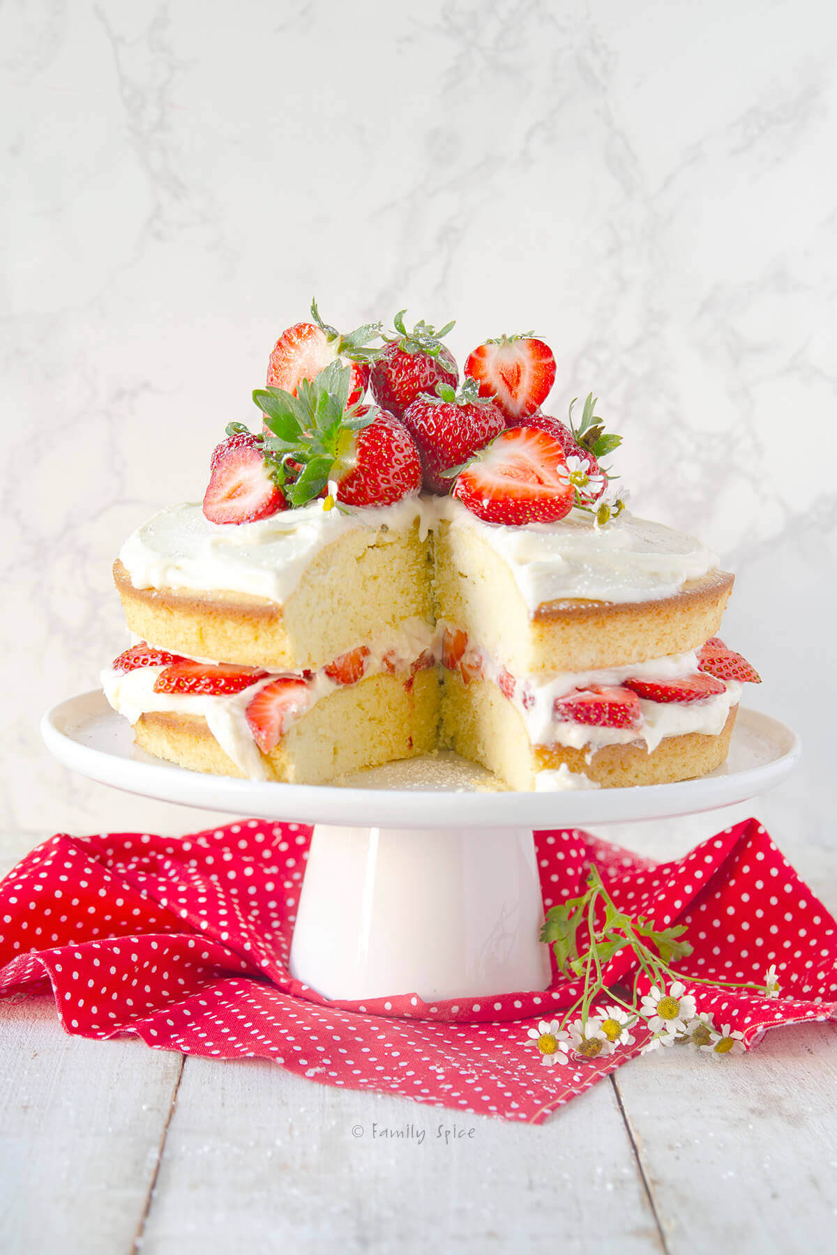 A vanilla cake frosted minimally with strawberry slices in between, with a slice cut out and topped with more fresh strawberries and chamomile flowers