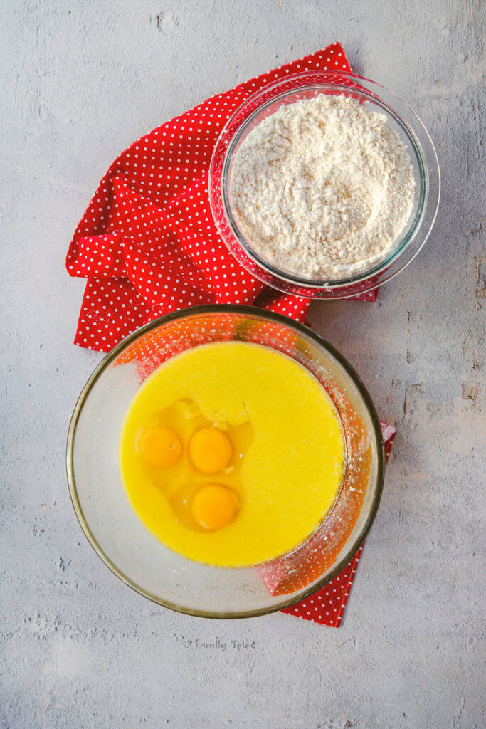 Adding eggs to a mixing bowl with olive oil vanilla cake batter in it and a bowl of flour mix next to it