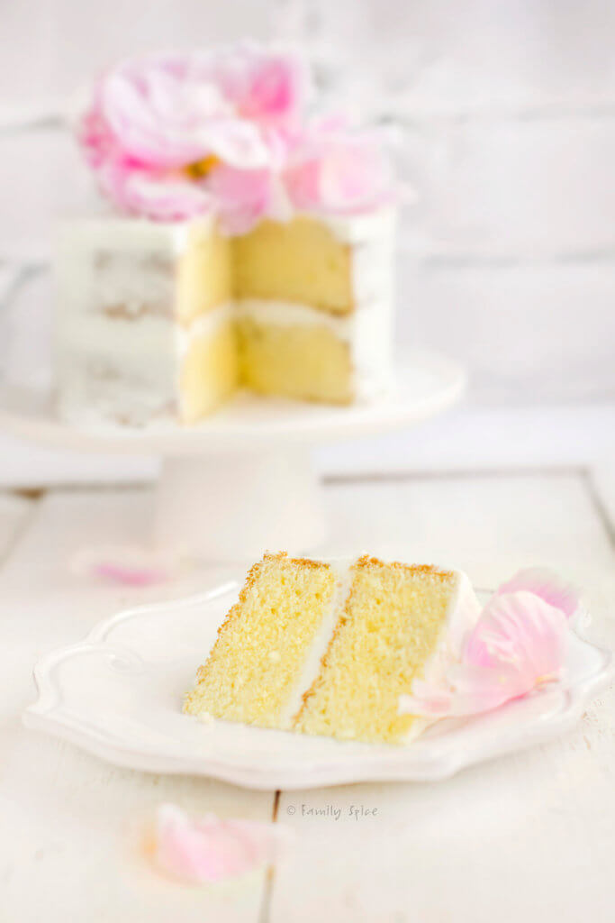 Closeup of a slice of vanilla cake with a 2 layer 6-inch round cake frosted minimally and topped with a large pink peony behind it