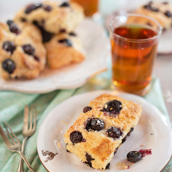 Closeup of a blueberry sour cream scone on a white plate with tea and more scones behind it