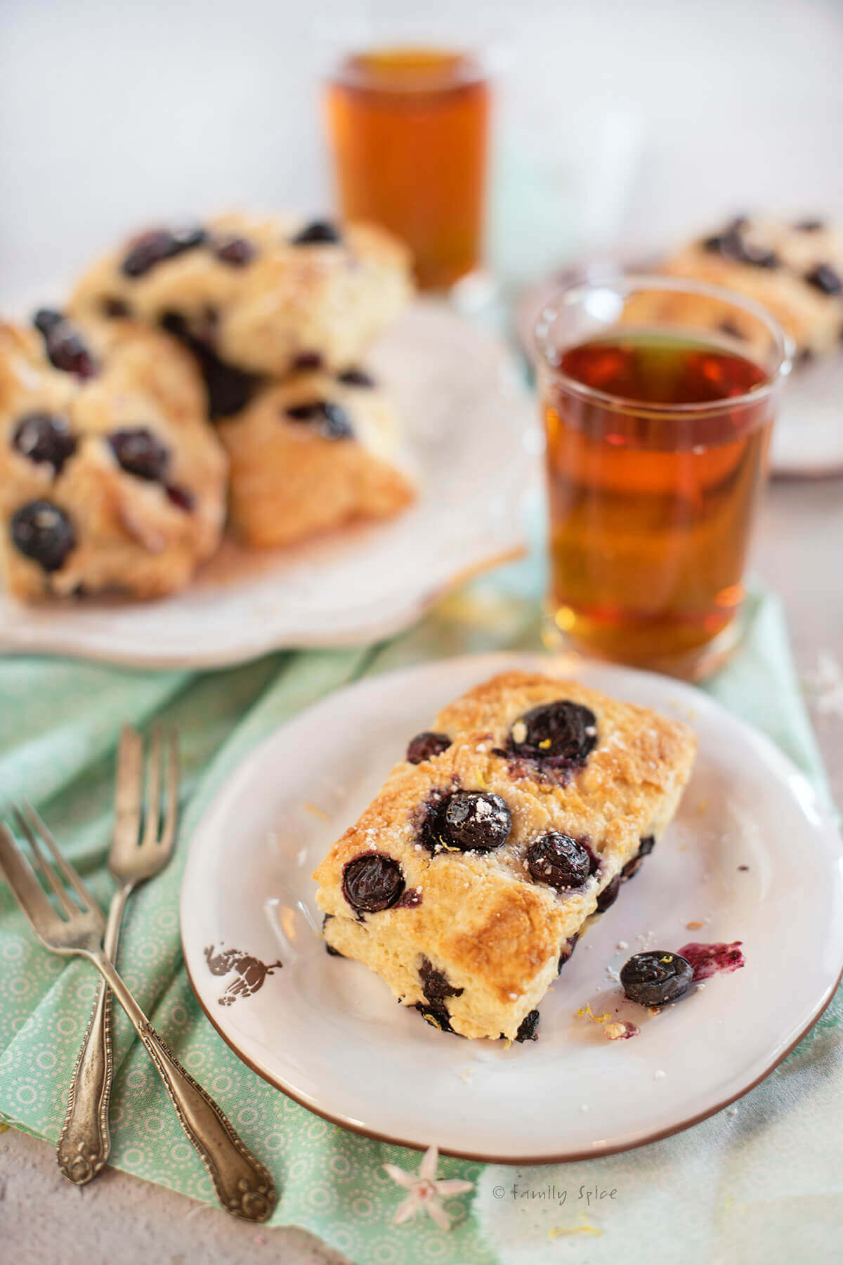 Closeup of a blueberry sour cream scone on a white plate with tea and more scones behind it
