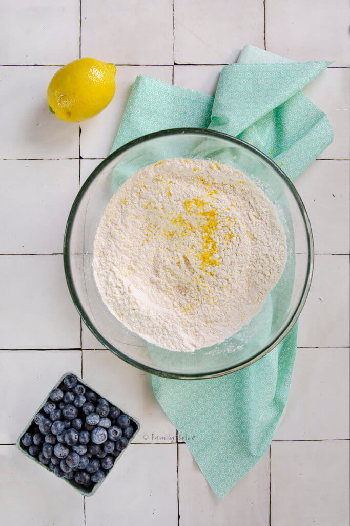 A mixing bowl with flour mixture and grated zest in it with a lemon and blueberries next to it