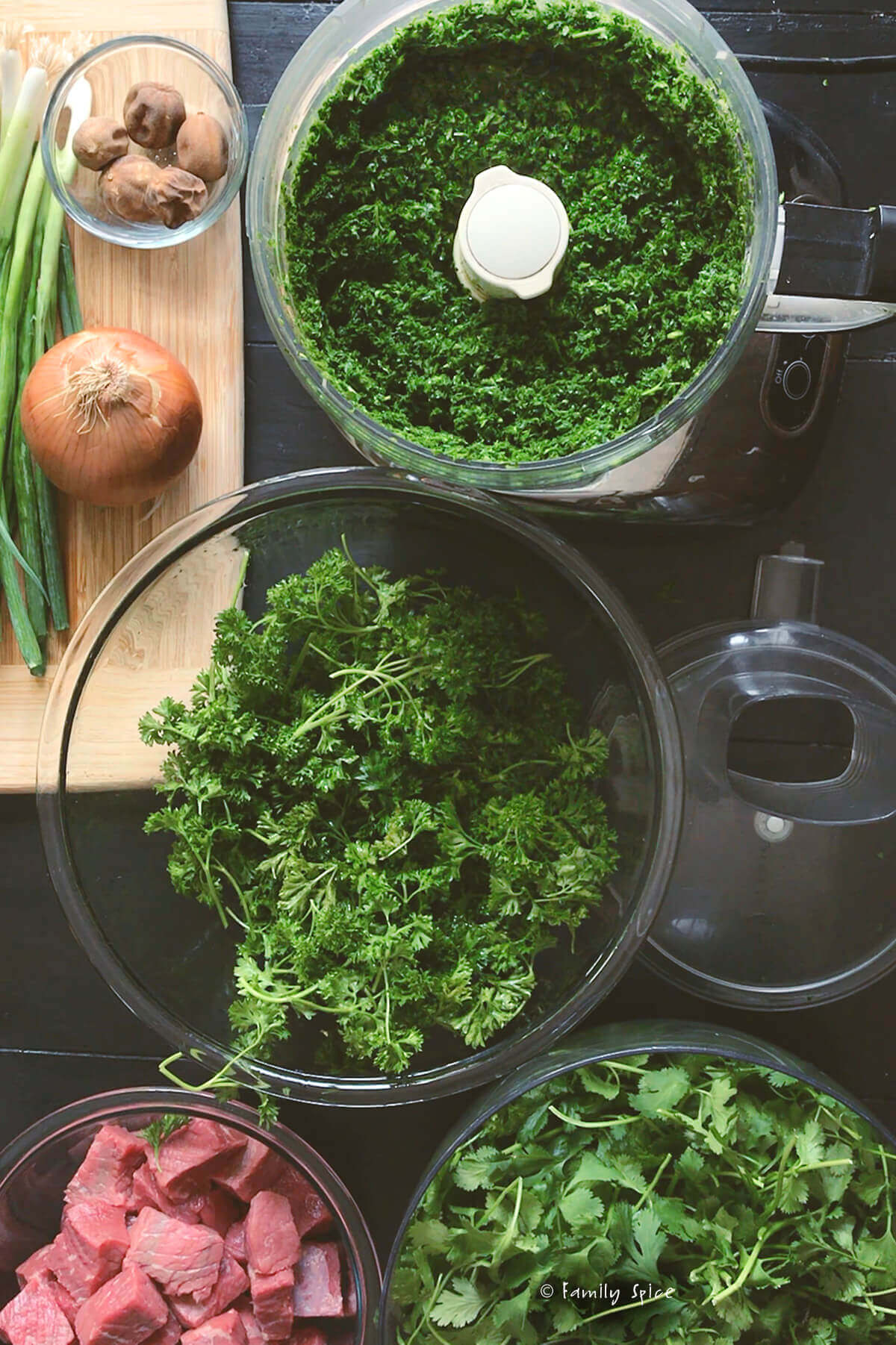 Parsley chopped in a food processor with bowls with fresh parsley and cilantro and other ingredients next to it