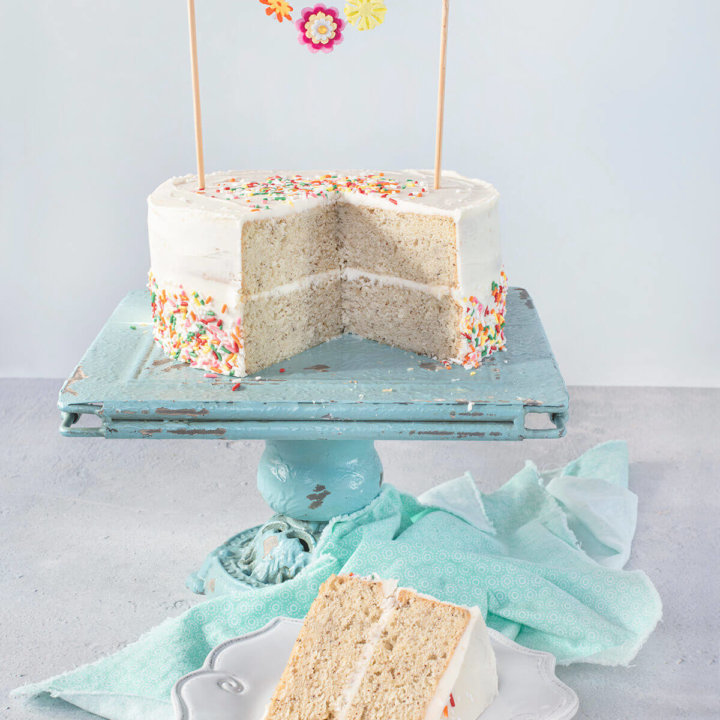 A vegan birthday cake covered with colorful sprinkles cut open on a turquoise blue cake stand with a slice of cake on a white plate next to it