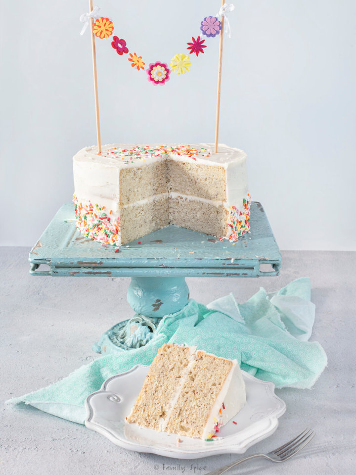 A vegan birthday cake covered with colorful sprinkles cut open on a turquoise blue cake stand with a slice of cake on a white plate next to it