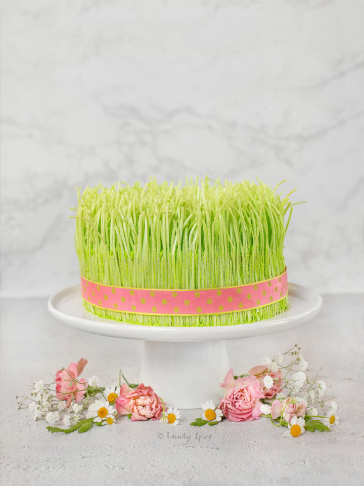A Spring Cake resembling sprouts with a pink ribbon wrapped around it on a cake stand with pink and white flowers around it