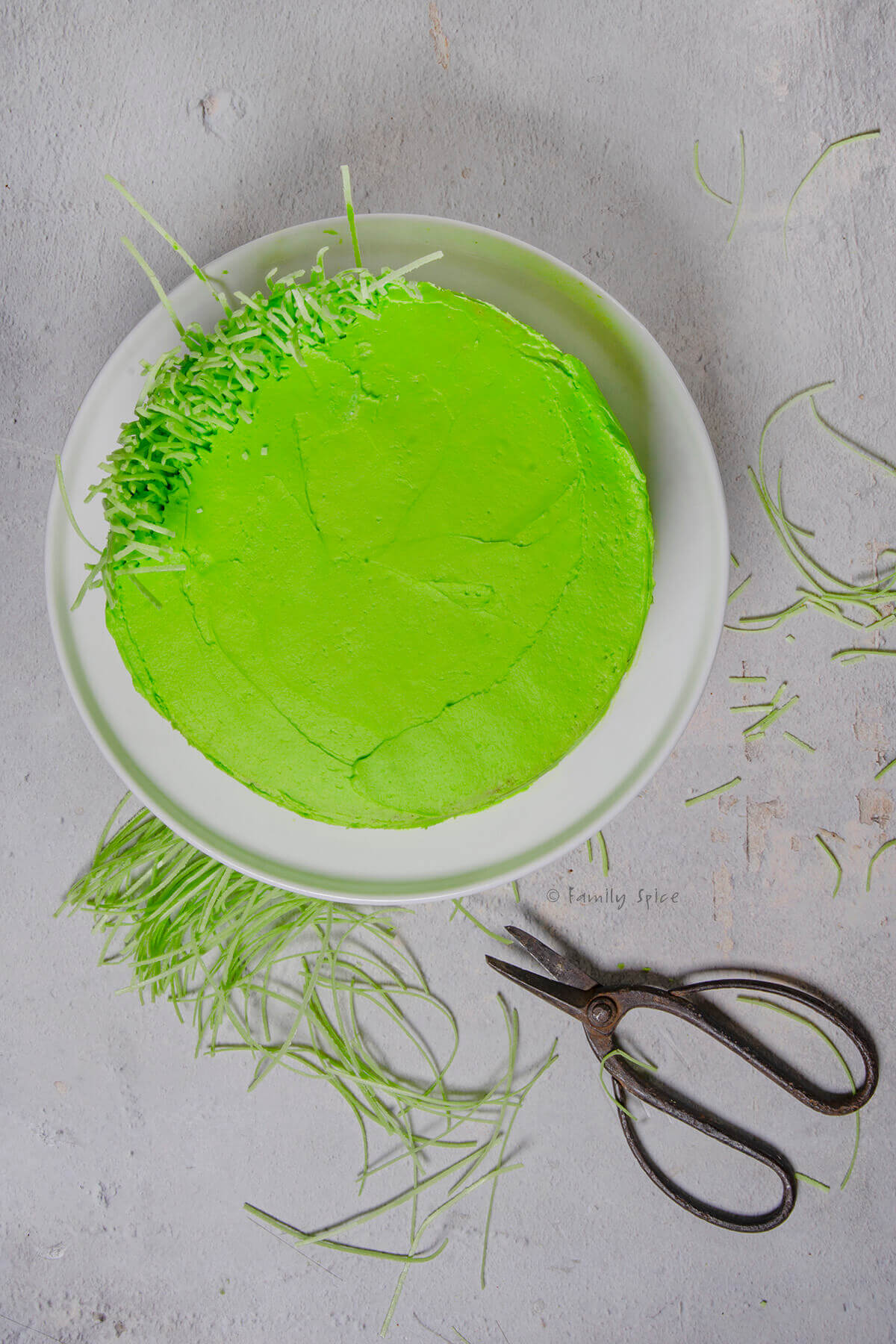 Top view of adding edible candy grass to a green frosted cake along its sides and on top with scissors next to it
