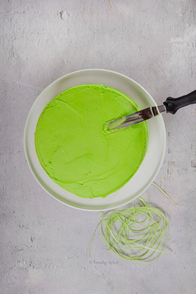 Frosting a cake with bright green frosting sitting on a white cake stand
