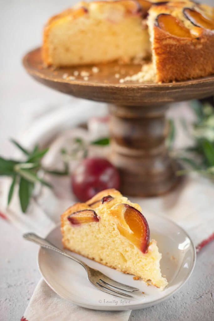 Closeup of a slice of yogurt plum cake with the rest of the cake on a wood cake stand behind it