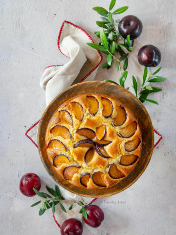 Top view of a plum cake on wood cake stand with olive branches and plums next to it