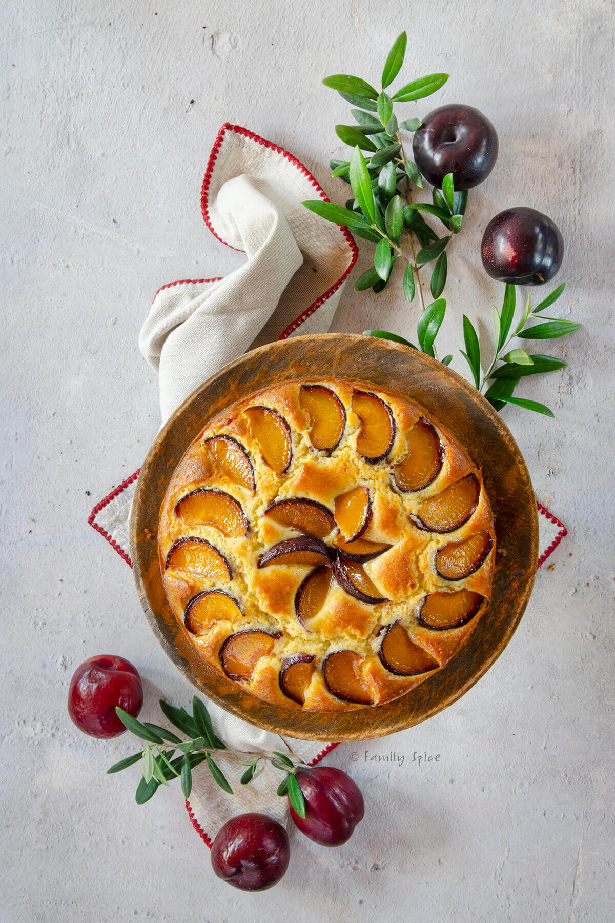 Top view of a plum cake on wood cake stand with olive branches and plums next to it