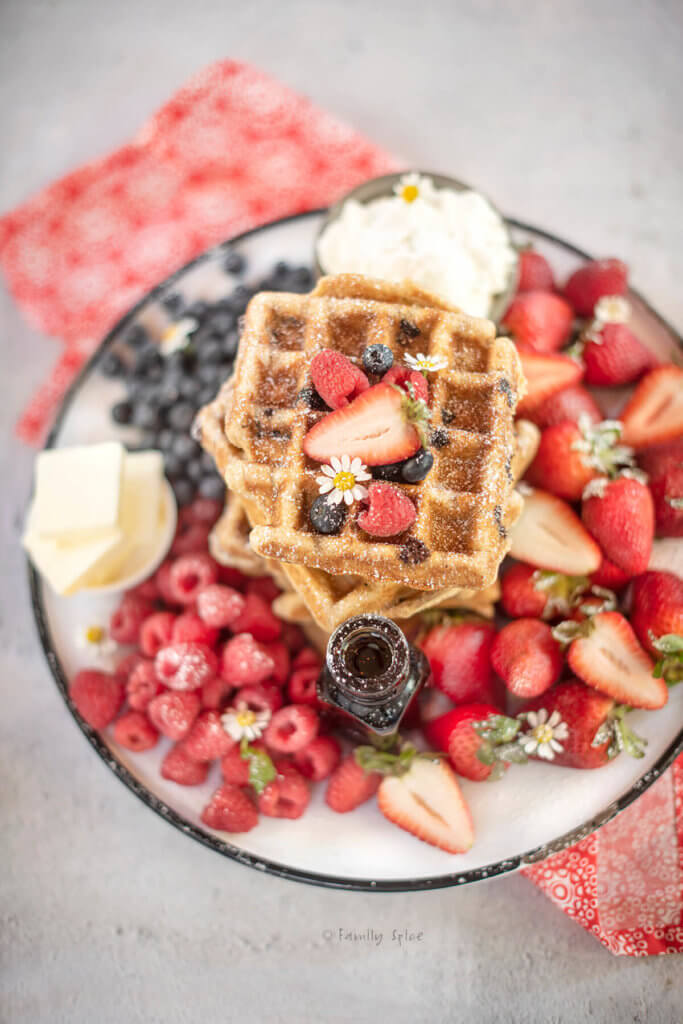 Closeup of the top waffle on a round enamel platter with more waffles, fresh berries, syrup, whipped cream and butter